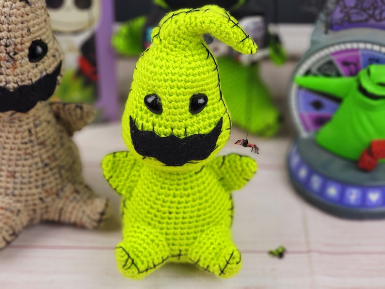 Puede incluir: Un juguete de amigurumi de crochet verde con forma de fantasma con una boca cosida negra y un bigote negro. El juguete tiene ojos negros y un gorro de crochet verde.
