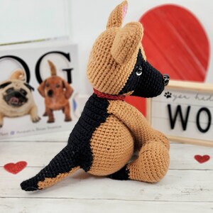 May include: A crocheted German Shepherd dog plush toy with brown and black fur, sitting on a white wooden surface. The dog is wearing a red collar and has a black nose and brown eyes. The toy is surrounded by red glitter hearts.