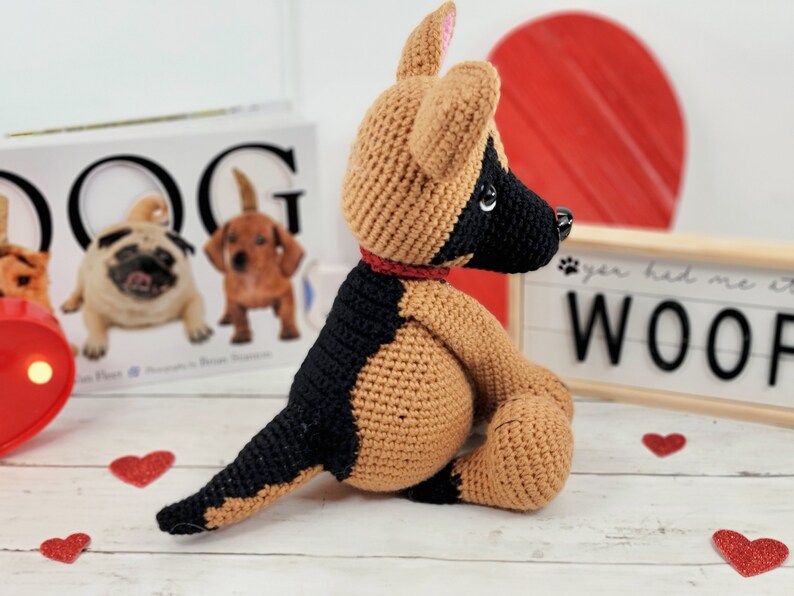 May include: A crocheted German Shepherd dog plush toy with brown and black fur, sitting on a white wooden surface. The dog is wearing a red collar and has a black nose and brown eyes. The toy is surrounded by red glitter hearts.