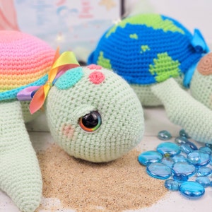 Puede incluir: Dos juguetes de tortuga marina de crochet. Uno es verde con rayas de arco&iacute;ris y un lazo rosa. El otro es azul con rayas verdes. Ambos tienen ojos saltones y est&aacute;n sentados sobre una superficie arenosa con cuentas de vidrio azules.