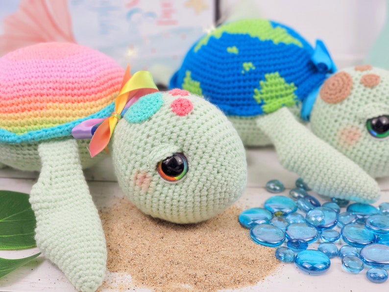 Puede incluir: Dos juguetes de tortuga marina de crochet. Uno es verde con rayas de arco&iacute;ris y un lazo rosa. El otro es azul con rayas verdes. Ambos tienen ojos saltones y est&aacute;n sentados sobre una superficie arenosa con cuentas de vidrio azules.