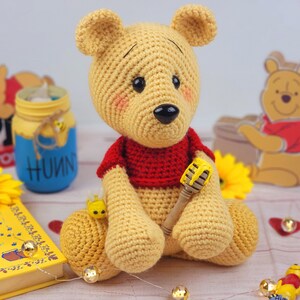 May include: A crocheted Winnie the Pooh bear toy wearing a red shirt and holding a yellow honey pot. The bear is sitting on a wooden surface with a book, a jar of honey, and a wooden Winnie the Pooh figurine in the background.