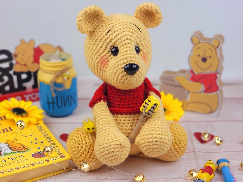 May include: A crocheted Winnie the Pooh bear toy wearing a red shirt and holding a yellow honey pot. The bear is sitting on a wooden surface with a book, a jar of honey, and a wooden Winnie the Pooh figurine in the background.