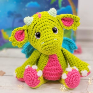 Puede incluir: Un drag&oacute;n de crochet verde con orejas, alas y pies rosas. El drag&oacute;n tiene ojos grandes y negros y una nariz rosa.
