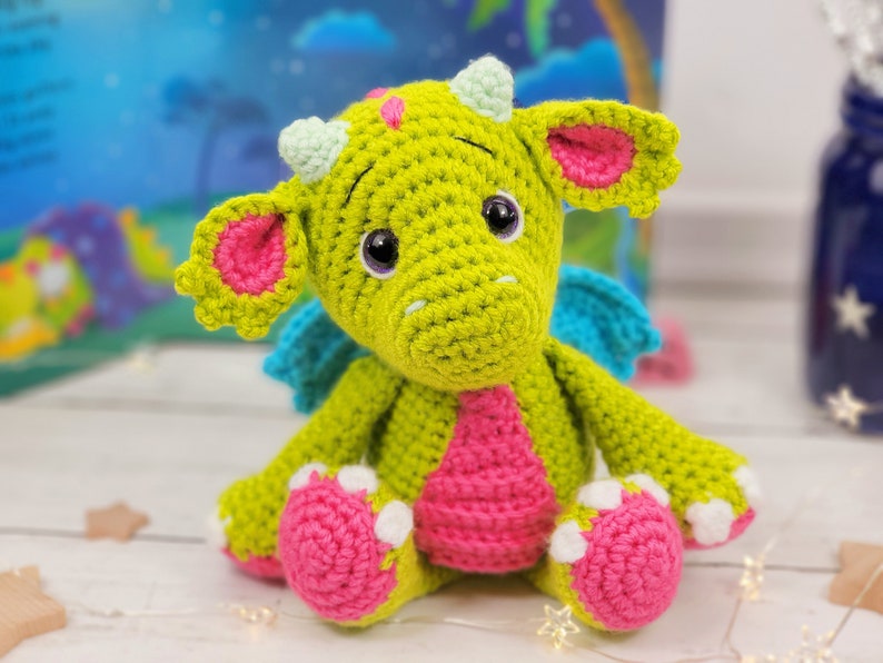 Puede incluir: Un drag&oacute;n de crochet verde con orejas, alas y pies rosas. El drag&oacute;n tiene ojos grandes y negros y una nariz rosa.