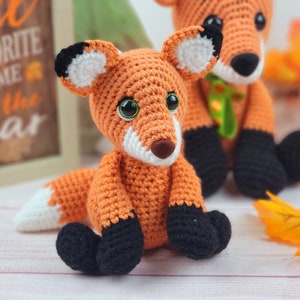 May include: Two crocheted fox plushies, one orange and white with black accents, the other orange with black accents, sitting on a white surface with fall leaves.