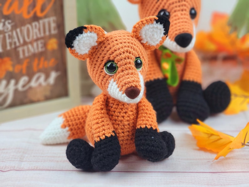 May include: Two crocheted fox plushies, one orange and white with black accents, the other orange with black accents, sitting on a white surface with fall leaves.