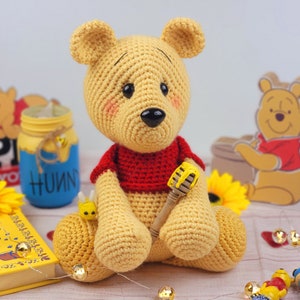 K&ouml;nnte beinhalten: Ein geh&auml;keltes Winnie-the-Pooh-B&auml;ren-Spielzeug mit einem roten Shirt und einem Honigl&ouml;ffel. Der B&auml;r sitzt auf einem Tisch mit einem Buch, einem Honigglas und Lichterketten.