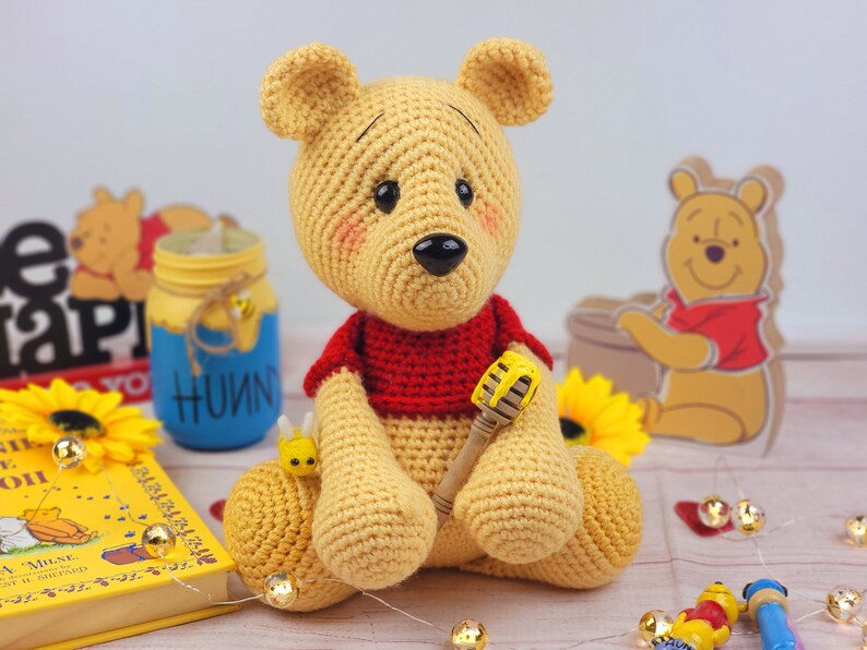 K&ouml;nnte beinhalten: Ein geh&auml;keltes Winnie-the-Pooh-B&auml;ren-Spielzeug mit einem roten Shirt und einem Honigl&ouml;ffel. Der B&auml;r sitzt auf einem Tisch mit einem Buch, einem Honigglas und Lichterketten.