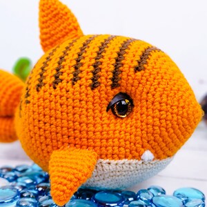 K&ouml;nnte beinhalten: Ein geh&auml;keltes orange-braunes Hai-Spielzeug mit wei&szlig;em Bauch und schwarzen Augen. Der Hai sitzt auf einer wei&szlig;en Oberfl&auml;che mit blauen Glassteinen.