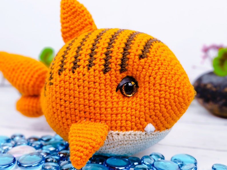 K&ouml;nnte beinhalten: Ein geh&auml;keltes orange-braunes Hai-Spielzeug mit wei&szlig;em Bauch und schwarzen Augen. Der Hai sitzt auf einer wei&szlig;en Oberfl&auml;che mit blauen Glassteinen.