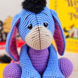 May include: A crocheted blue and purple Eeyore plush toy with a black fuzzy mane. The toy is sitting on a white surface.