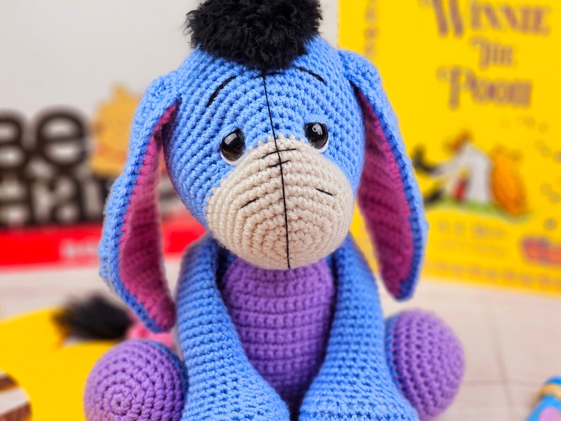 May include: A crocheted blue and purple Eeyore plush toy with a black fuzzy mane. The toy is sitting on a white surface.