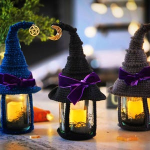 Pode incluir: Três lanternas de frasco de vidro cobertas com chapéus de bruxa de crochê em azul, preto e cinza. Cada chapéu apresenta um laço de veludo roxo e um pingente dourado, incluindo um pentáculo e uma lua crescente. Dentro de cada frasco há uma vela LED amarela.
