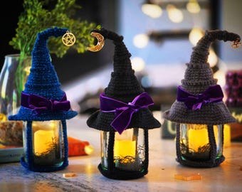 May include: Three glass jar lanterns topped with crocheted witch hats in blue, black, and gray. Each hat features a purple velvet ribbon bow and a gold-toned charm, including a pentacle and a crescent moon. Inside each jar sits a yellow LED candle.
