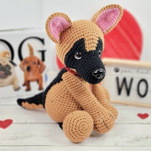 May include: A crocheted German Shepherd puppy toy with brown and black fur, pink ears, and a red collar. The toy is sitting on a white wooden surface with red hearts scattered around it.
