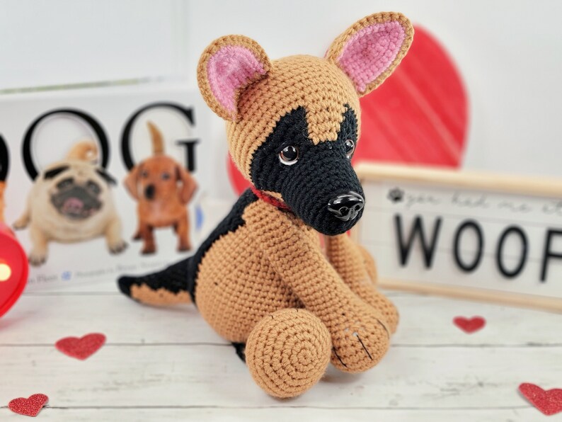 May include: A crocheted German Shepherd puppy toy with brown and black fur, pink ears, and a red collar. The toy is sitting on a white wooden surface with red hearts scattered around it.