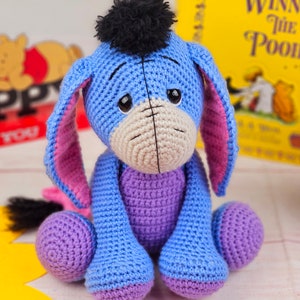 May include: A crocheted stuffed toy donkey with blue fur, pink ears, and a black pom-pom on its head. The donkey is sitting on a white surface with a book in the background.