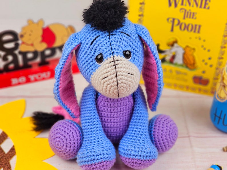 May include: A crocheted stuffed toy donkey with blue fur, pink ears, and a black pom-pom on its head. The donkey is sitting on a white surface with a book in the background.