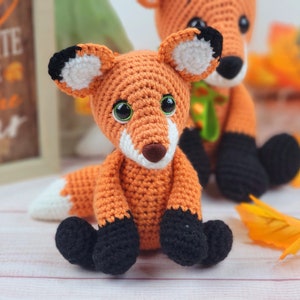 May include: Two crocheted fox plushies, one orange and white with black eyes and nose, the other orange with black eyes and nose, sitting on a white surface with fall leaves.