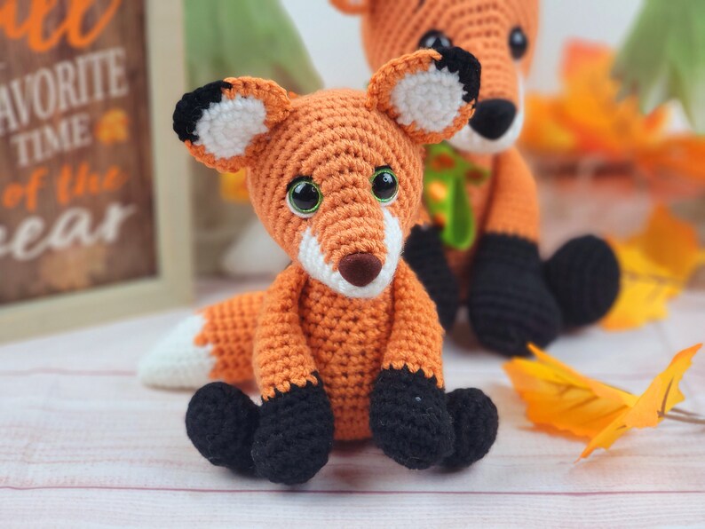 May include: Two crocheted fox plushies, one orange and white with black eyes and nose, the other orange with black eyes and nose, sitting on a white surface with fall leaves.