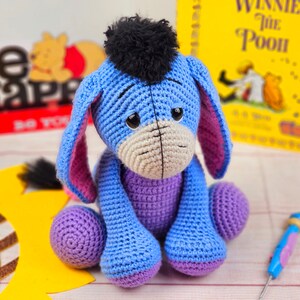 May include: A blue crocheted Eeyore plush toy with purple accents and a black yarn mane. The toy is sitting on a wooden surface with a sunflower and a book in the background.