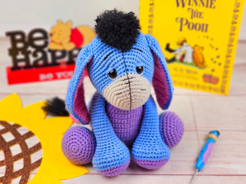 May include: A blue crocheted Eeyore plush toy with purple accents and a black yarn mane. The toy is sitting on a wooden surface with a sunflower and a book in the background.