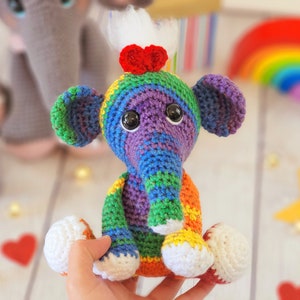 May include: A colorful crocheted elephant toy with a rainbow pattern and white accents. The elephant has a red heart on its head and is sitting on a hand.