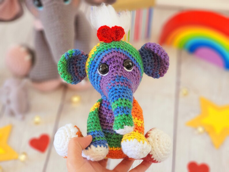 May include: A colorful crocheted elephant toy with a rainbow pattern and white accents. The elephant has a red heart on its head and is sitting on a hand.