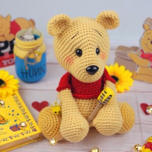 K&ouml;nnte beinhalten: Ein geh&auml;keltes Winnie Puuh Pl&uuml;schtier, das ein rotes Hemd tr&auml;gt und einen Honigtopf h&auml;lt. Das Spielzeug sitzt auf einem Tisch mit einem Winnie Puuh Buch und anderen Dekorationen.