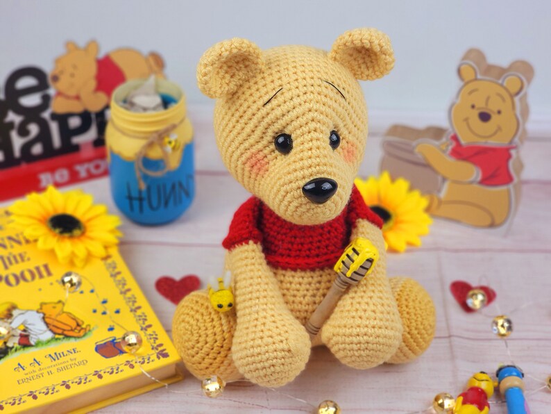 K&ouml;nnte beinhalten: Ein geh&auml;keltes Winnie Puuh Pl&uuml;schtier, das ein rotes Hemd tr&auml;gt und einen Honigtopf h&auml;lt. Das Spielzeug sitzt auf einem Tisch mit einem Winnie Puuh Buch und anderen Dekorationen.