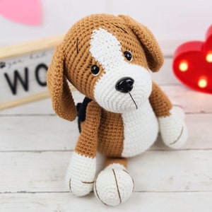 May include: A crocheted brown and white dog toy with black eyes and a black nose. The toy is sitting on a white wooden surface.