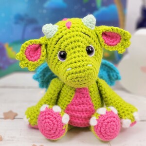 Puede incluir: Un drag&oacute;n de crochet verde con detalles en rosa y azul. El drag&oacute;n tiene ojos grandes y una expresi&oacute;n amigable.