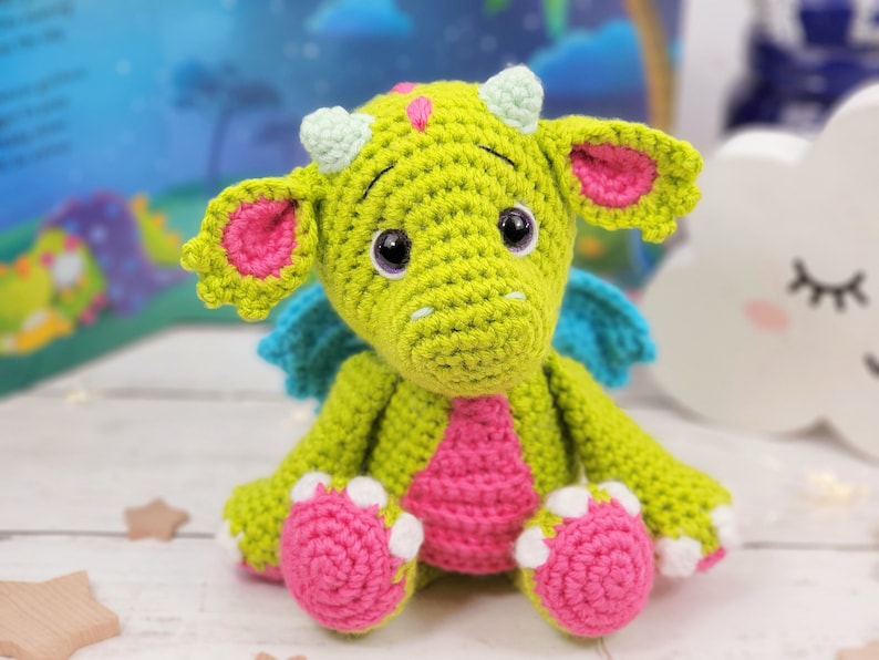 Puede incluir: Un drag&oacute;n de crochet verde con detalles en rosa y azul. El drag&oacute;n tiene ojos grandes y una expresi&oacute;n amigable.