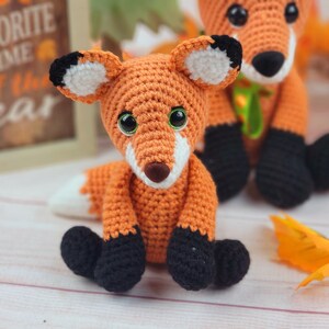 May include: Two crocheted fox plushies, one orange with black feet and a white patch on its forehead, the other orange with black feet and a green ribbon around its neck. The foxes are sitting on a white wooden surface with fall leaves.