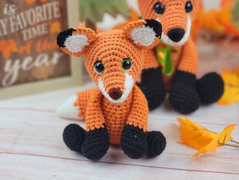 May include: Two crocheted fox plushies, one orange with black feet and a white patch on its forehead, the other orange with black feet and a green ribbon around its neck. The foxes are sitting on a white wooden surface with fall leaves.