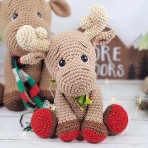 May include: Two crocheted reindeer toys, one brown with a red and green scarf and the other brown with red booties. The reindeer are sitting on a white surface with a wooden sign that says "More Doors" in the background.