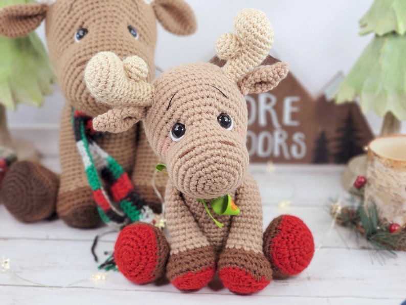 May include: Two crocheted reindeer toys, one brown with a red and green scarf and the other brown with red booties. The reindeer are sitting on a white surface with a wooden sign that says "More Doors" in the background.