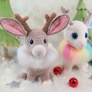 May include: Two crocheted reindeer-like creatures with large eyes and antlers. One is tan with a white fluffy collar, the other is white with a rainbow tutu. Christmas ornaments and a decorative tree are in the background.