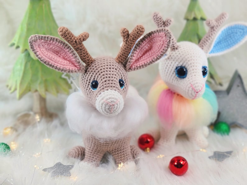 May include: Two crocheted reindeer-like creatures with large eyes and antlers. One is tan with a white fluffy collar, the other is white with a rainbow tutu. Christmas ornaments and a decorative tree are in the background.
