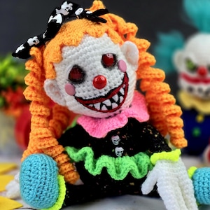 May include: A crocheted clown doll with orange hair, a black and white bow, and a creepy smile. The doll is wearing a black and green dress with a pink collar and white gloves. The doll is sitting on a yellow surface.