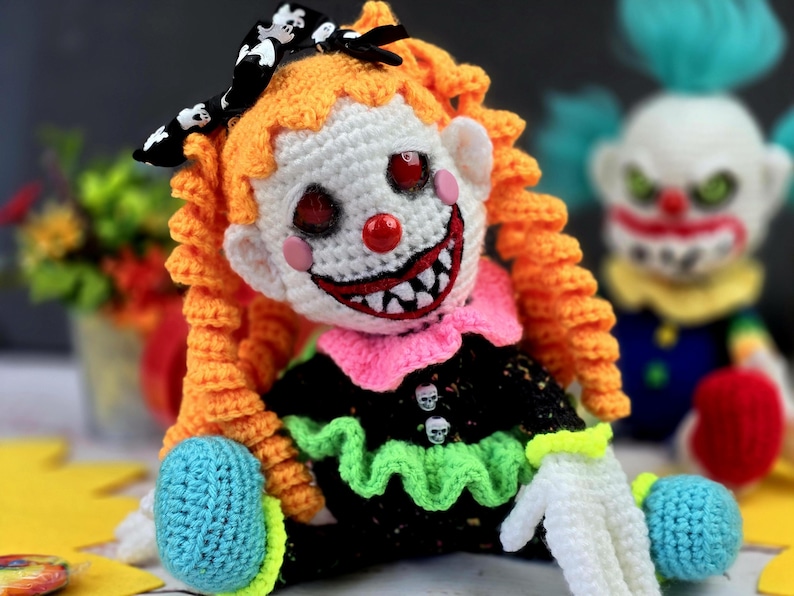 May include: A crocheted clown doll with orange hair, a black and white bow, and a creepy smile. The doll is wearing a black and green dress with a pink collar and white gloves. The doll is sitting on a yellow surface.