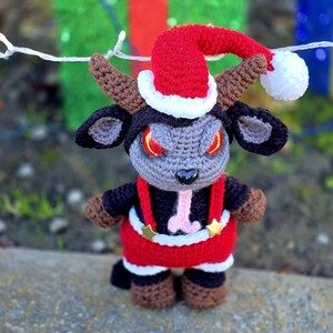 Puede incluir: Un juguete de peluche de crochet con forma de cabra que lleva un gorro de Pap&aacute; Noel rojo y blanco con un pomp&oacute;n blanco. La cabra tiene cuernos negros, ojos negros con pupilas rojas y una lengua rosa que sobresale. La cabra lleva un traje de Pap&aacute; Noel rojo y blanco con tirantes negros y estrellas doradas.