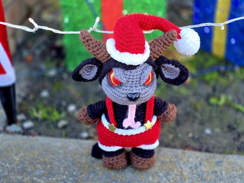 Puede incluir: Un juguete de peluche de crochet con forma de cabra que lleva un gorro de Pap&aacute; Noel rojo y blanco con un pomp&oacute;n blanco. La cabra tiene cuernos negros, ojos negros con pupilas rojas y una lengua rosa que sobresale. La cabra lleva un traje de Pap&aacute; Noel rojo y blanco con tirantes negros y estrellas doradas.