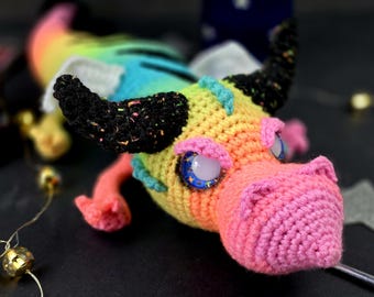 Könnte beinhalten: Ein farbenfrohes Häkel-Drachenspielzeug mit einer rosa Schnauze, schwarzen Hörnern und glitzernden blauen Augen. Der Drache ist mit einem Regenbogen aus Farben hergestellt, darunter Gelb, Orange, Rot, Rosa, Blau, Grün und Lila.