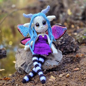 May include: A crocheted fairy doll with blue hair, purple dress, and black and white striped legs. The doll is sitting on a rock with wings spread out.
