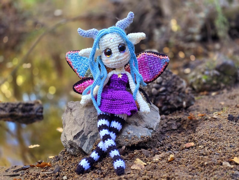 May include: A crocheted fairy doll with blue hair, purple dress, and black and white striped legs. The doll is sitting on a rock with wings spread out.