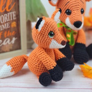 May include: Two crocheted fox plushies, one orange and one smaller orange with a white tip on its tail. The larger fox is sitting with its legs crossed and has a green ribbon around its neck. The smaller fox is sitting with its legs crossed and has black feet. The plushies are on a white wooden surface with fall leaves in the background.