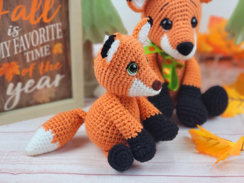 May include: Two crocheted fox plushies, one orange and one smaller orange with a white tip on its tail. The larger fox is sitting with its legs crossed and has a green ribbon around its neck. The smaller fox is sitting with its legs crossed and has black feet. The plushies are on a white wooden surface with fall leaves in the background.