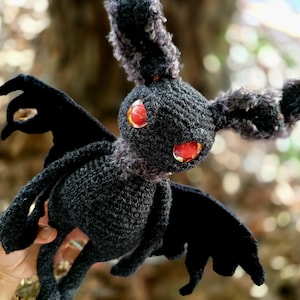Könnte beinhalten: Eine schwarze gehäkelte Fledermaus mit roten Augen. Die Fledermaus hat große Flügel und einen flauschigen Kopf.